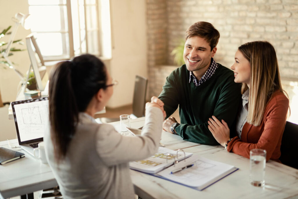 A Smiling Couple Sitting At A Desk, Shaking Hands With A Female Professional In An Office Setting.